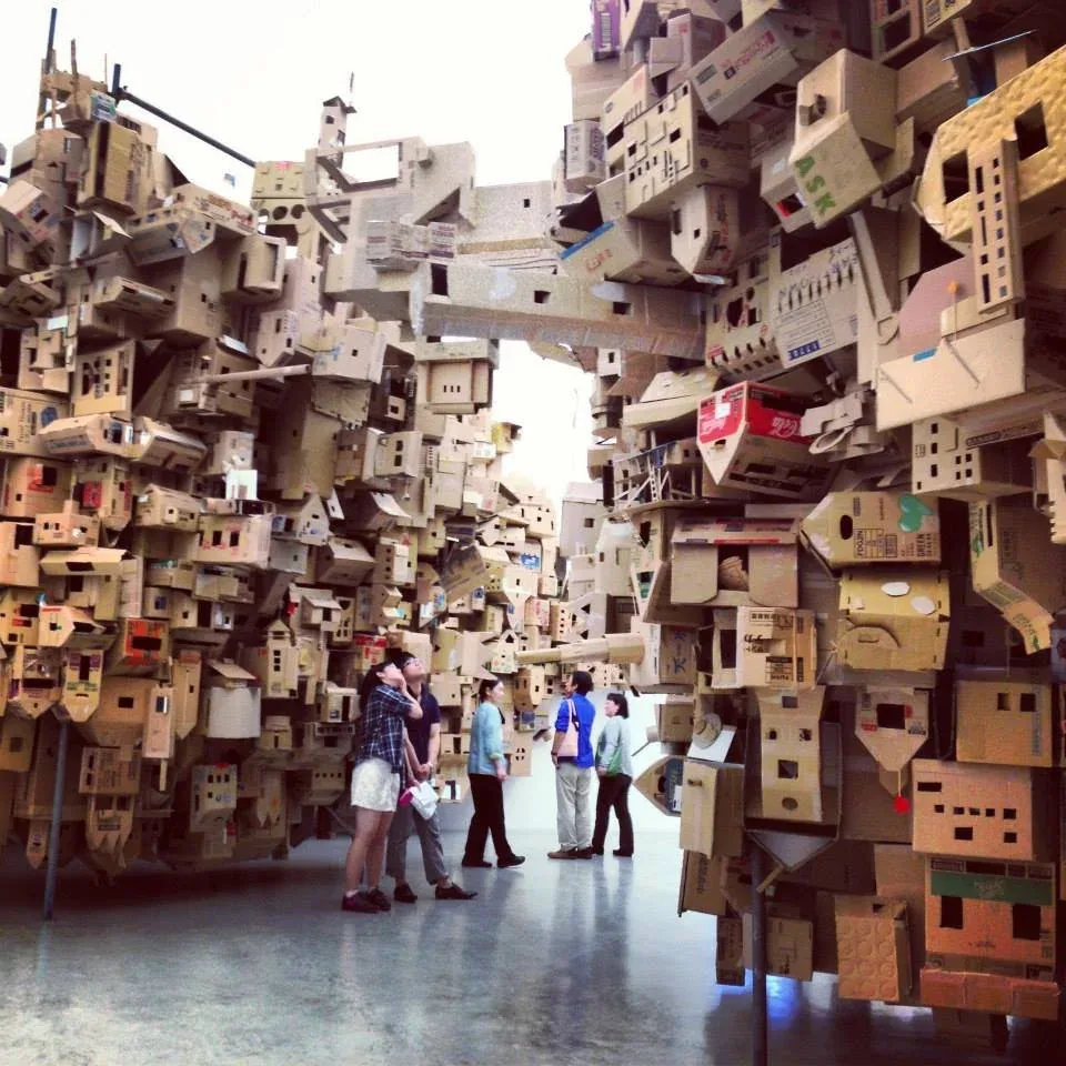 Visitors looking up at a towering installation built from cardboard boxes
